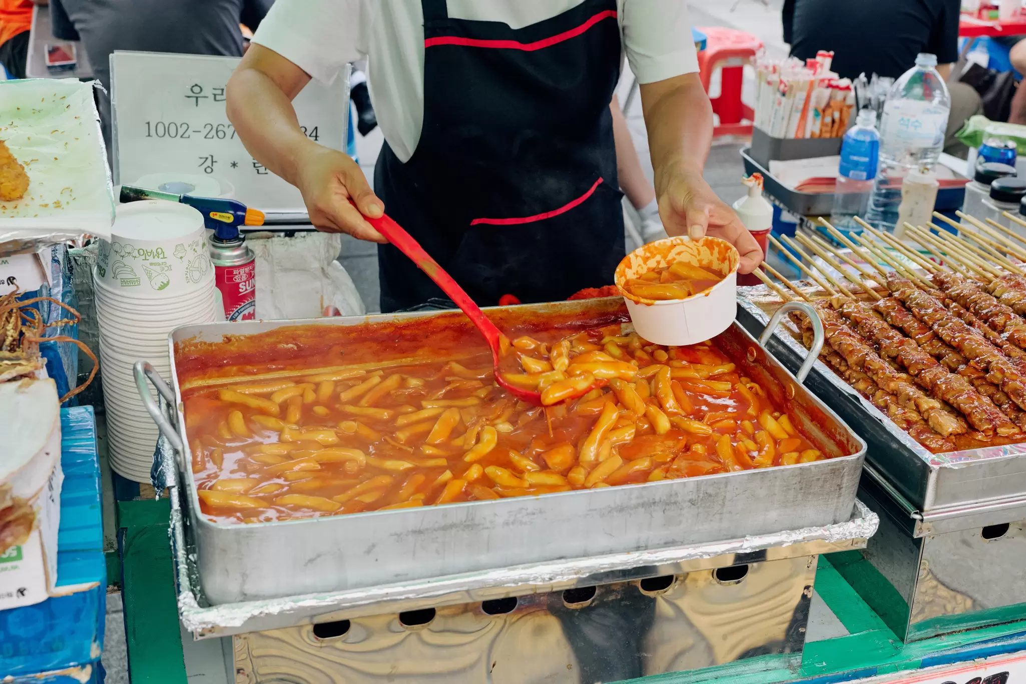 A person scoops a dish with red sauce and oblong dumplings into a container.