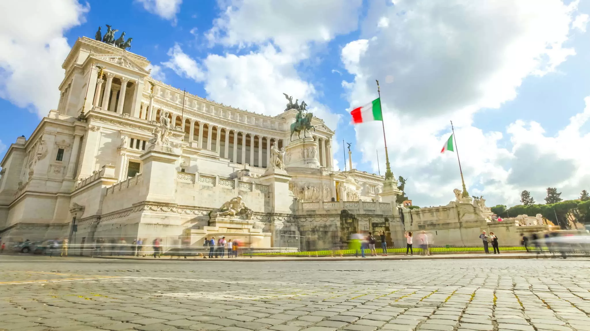 A massive white building surrounded by many statues with Italian flags flying outside