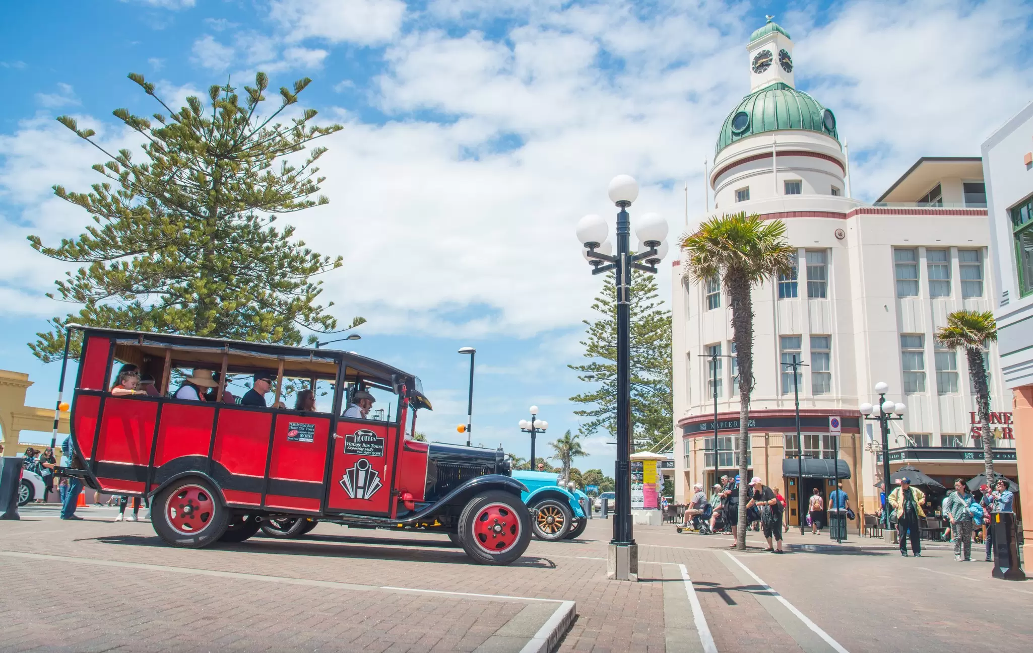 Vintage red open-sided tour bus passing an Art Deco building with a green domed clock tower in Napier, New Zealand