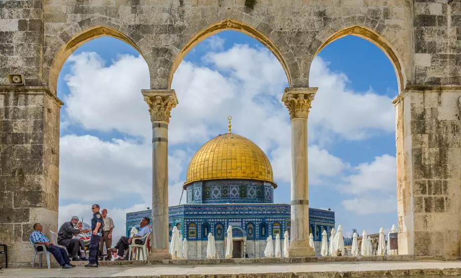 Dome of the Rock in Jerusalem's Old City, its gold dome and blue tiles framed by stone arches, with men seated in the shade