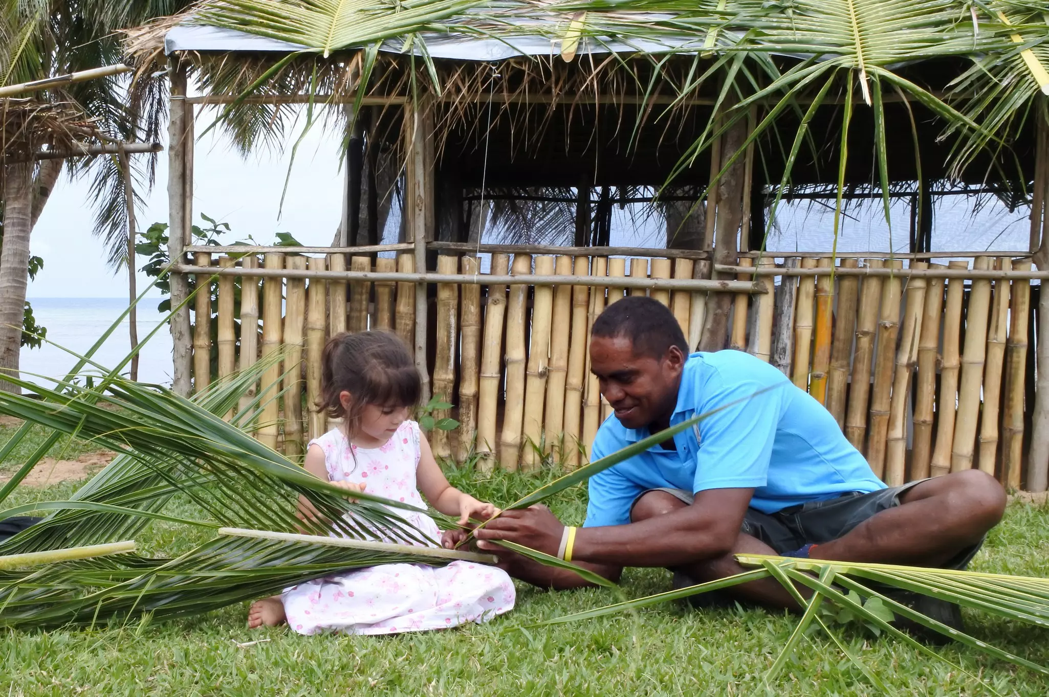 A man sits with a young girl, teaching her how to weave palm fronds on a lawn in front of a covered pavilion.
