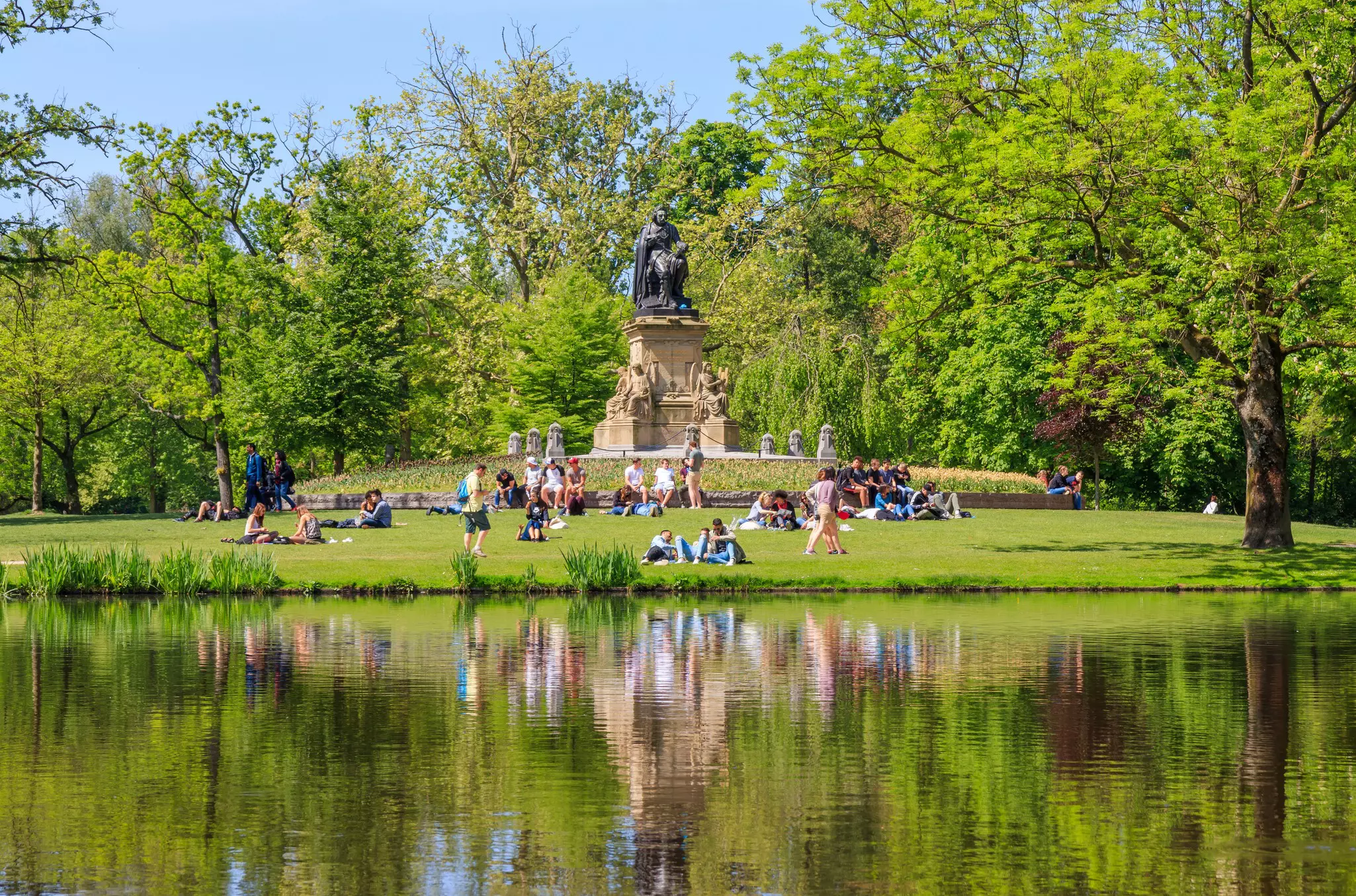 People sit and relax on the lawns near a lake in green parkland 