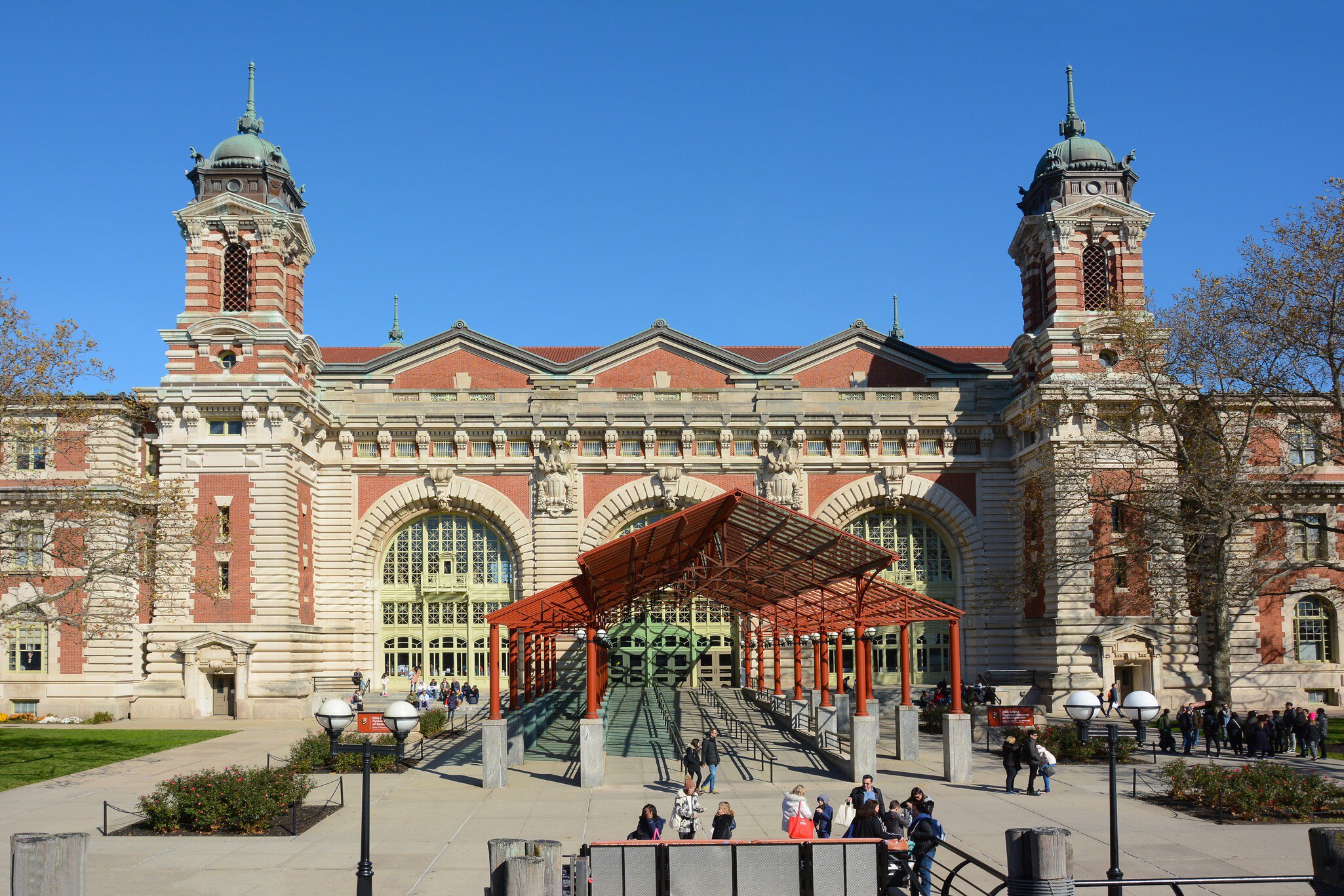 A red steel canopy shelters the glass entrance ramp below twin copper-domed towers; visitors cross the paved plaza.