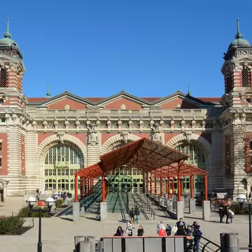 A red steel canopy shelters the glass entrance ramp below twin copper-domed towers; visitors cross the paved plaza.