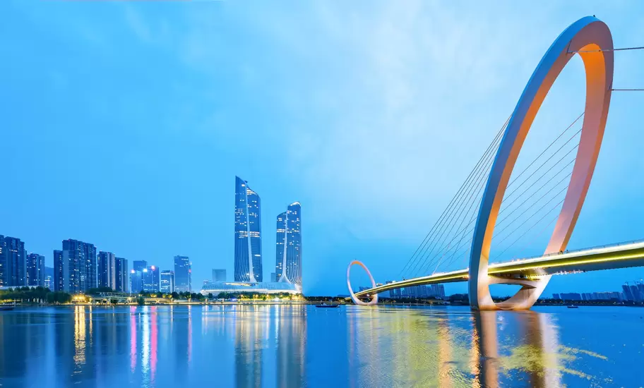 Nanjing Eye footbridge with tilted ring arches over the Yangtze at dusk, twin curved towers of the Hexi skyline behind