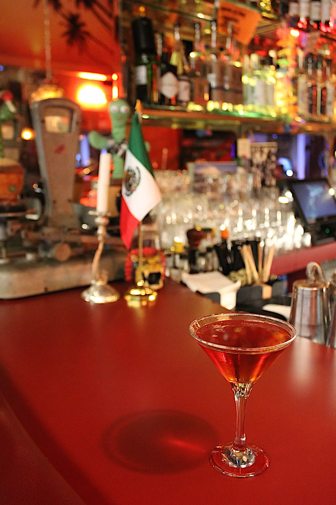 A red cocktail in a martini glass on a red counter in front of a Mexican flag and various bar paraphenalia