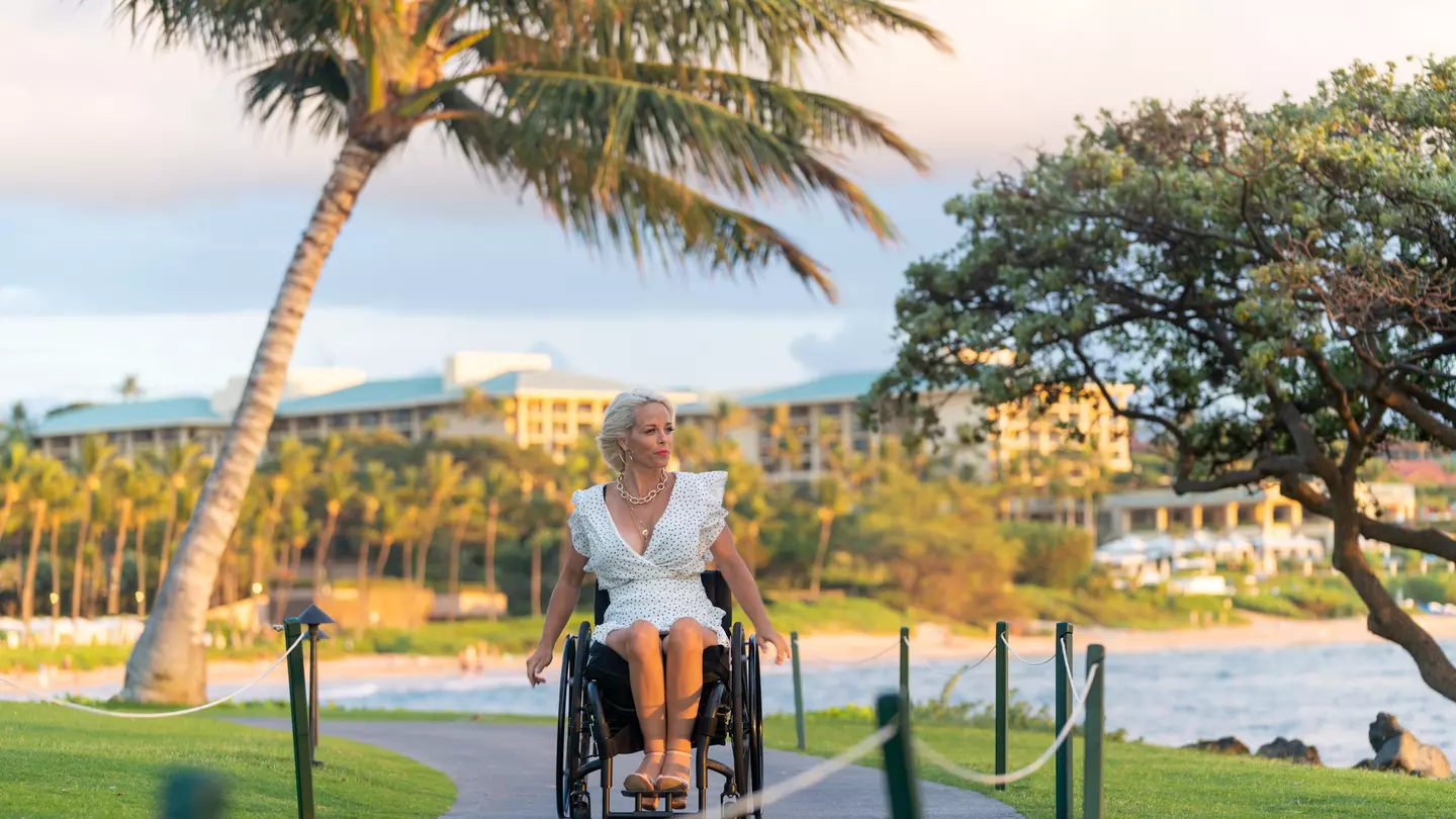 A white woman in a white dress wheels her wheelchair along a boardwalk in a tropical destination