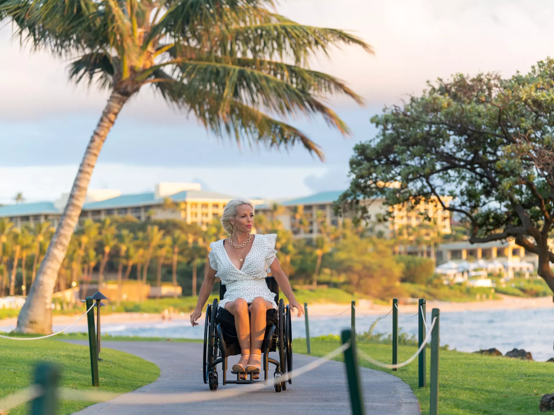 A white woman in a white dress wheels her wheelchair along a boardwalk in a tropical destination