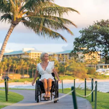 A white woman in a white dress wheels her wheelchair along a boardwalk in a tropical destination