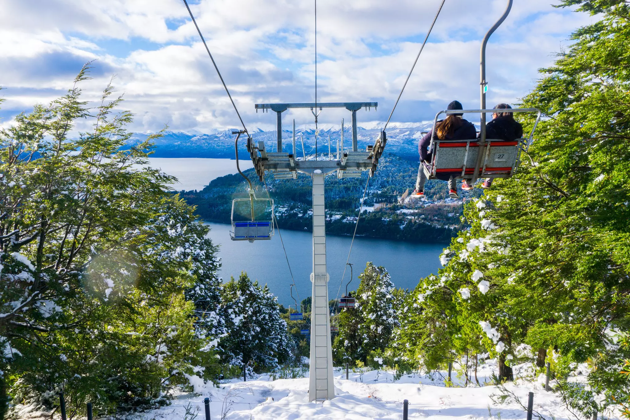 People ride a chair lift over a snowy hillside by a blue lake.