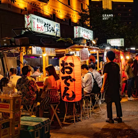 People eat at a food stall on a dark street in Japan with red laterns. 