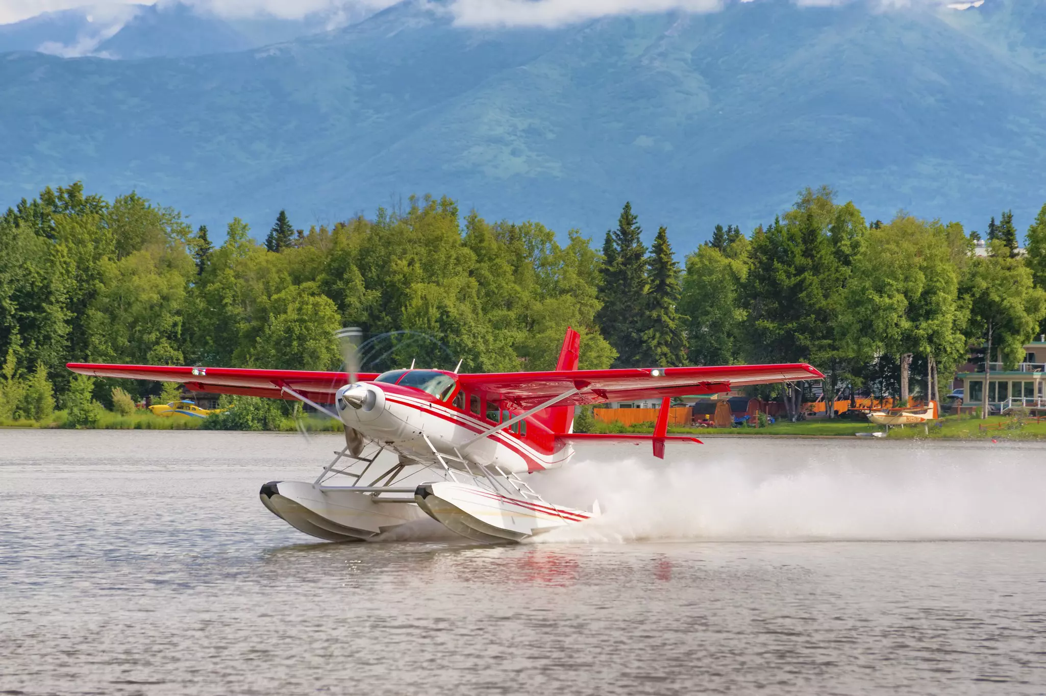 A red and white seaplane takes off on Lake Hood in Anchorage, Alaska, with trees behind.