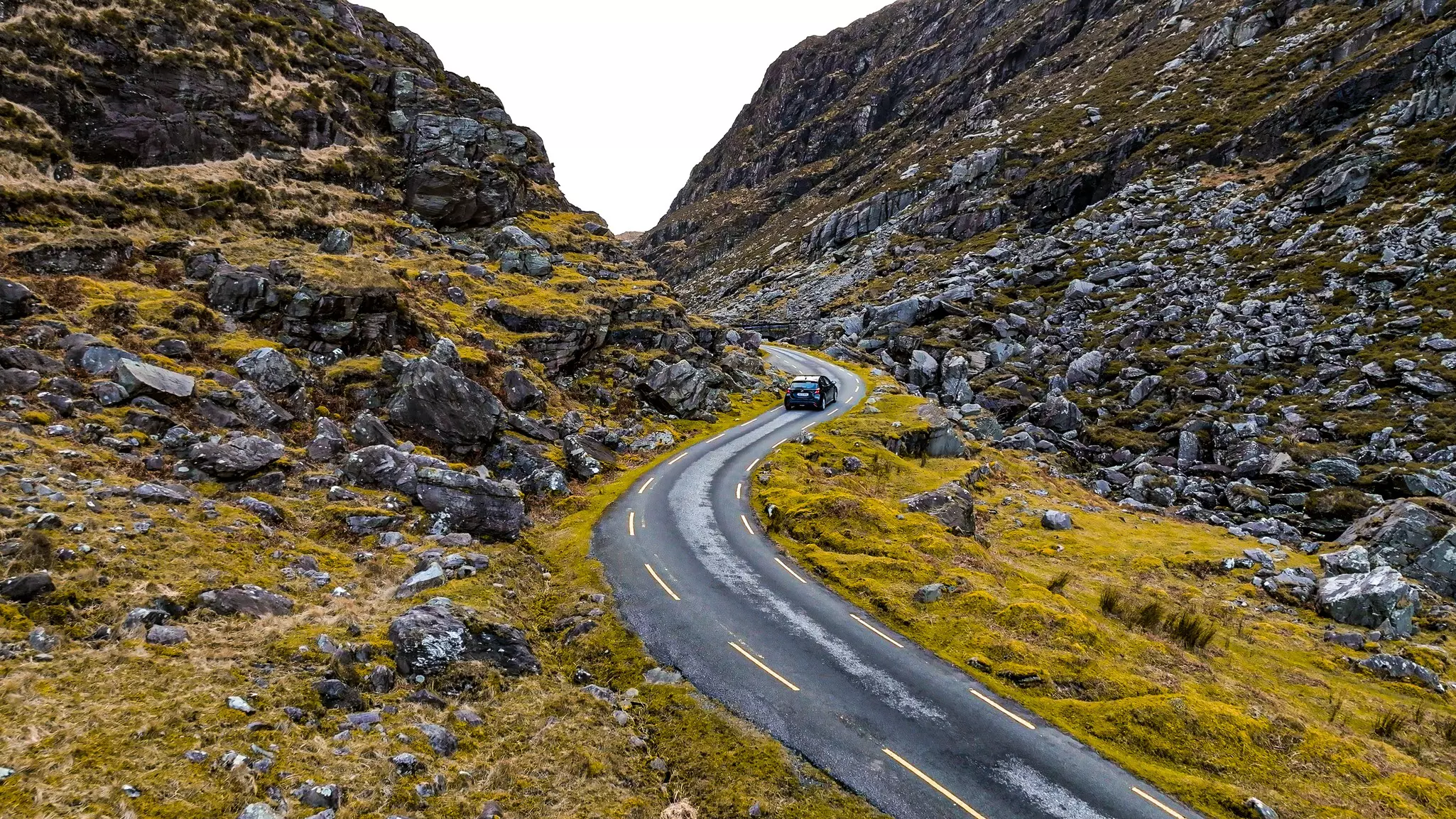 A car drives on a narrow blacktop road through a rocky landscape.