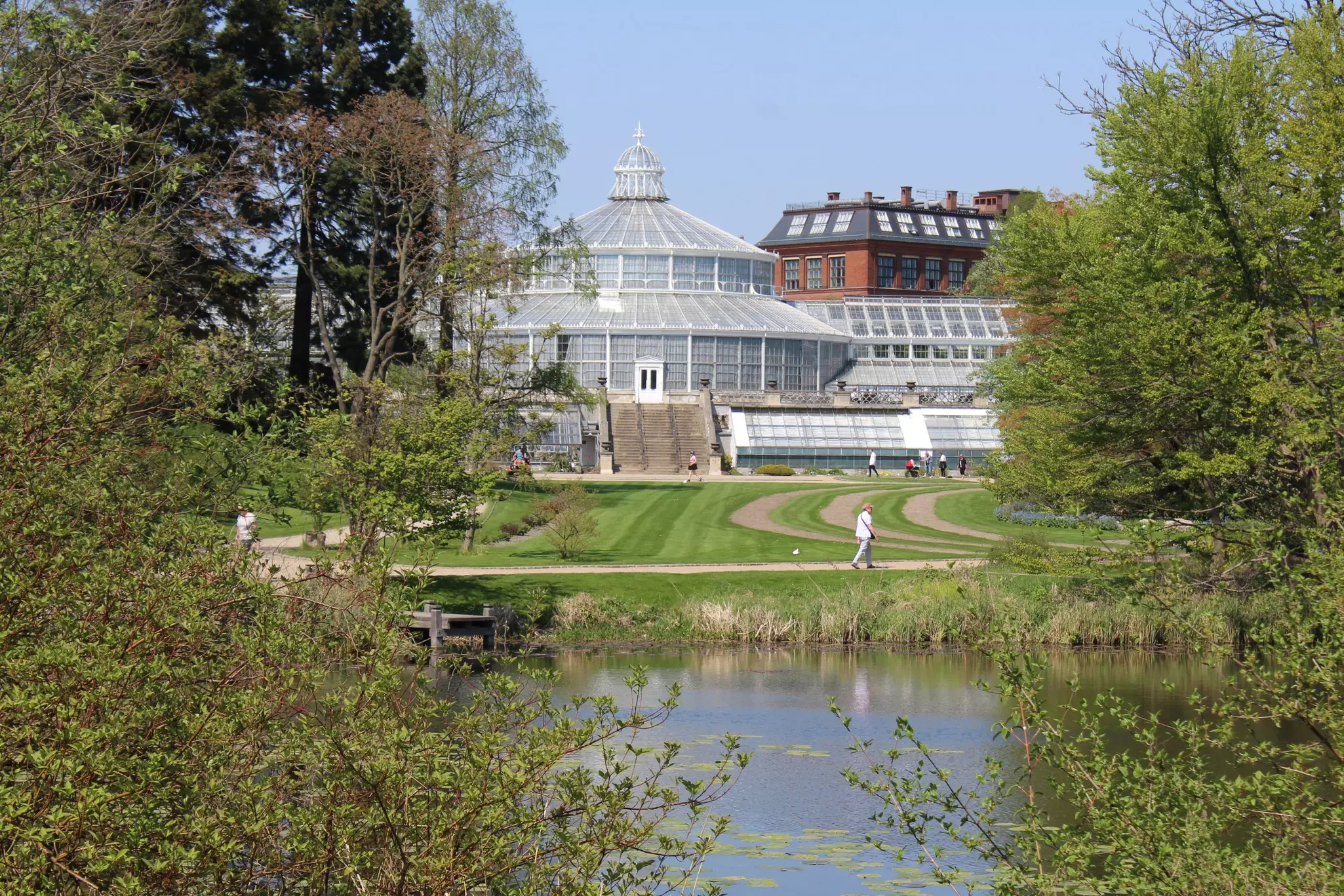 Tree branches in the foreground frame a large greenhouse reached by many stairs from a green lawn; people are walking on paths through the lawn, which borders a pond.