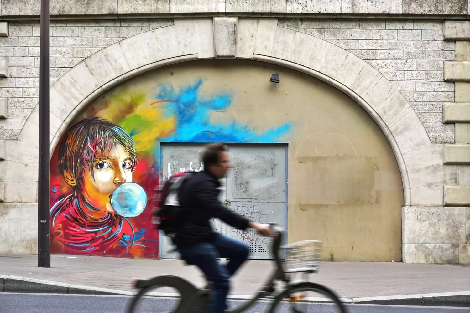 A blurred cyclist passing a colorful mural of a figure blowing a bubble.