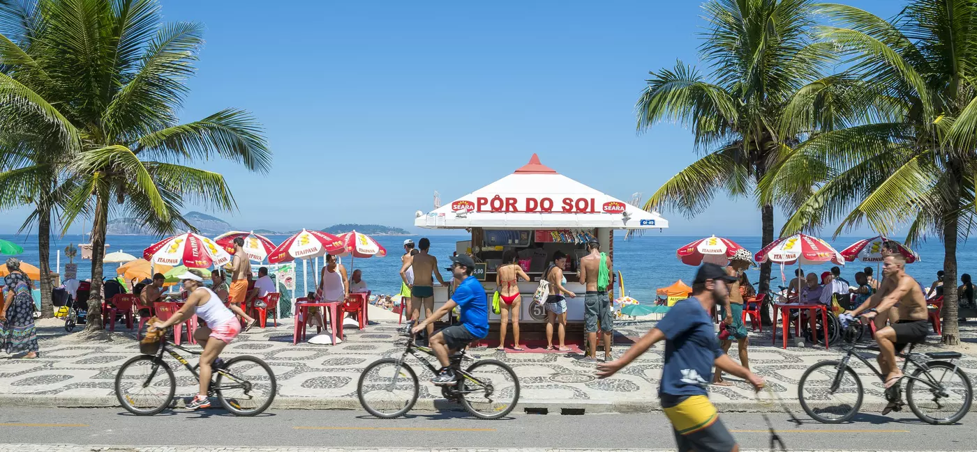  Cyclists and skateboarder pass a typical beach kiosk on the boardwalk at Ipanema Beach  