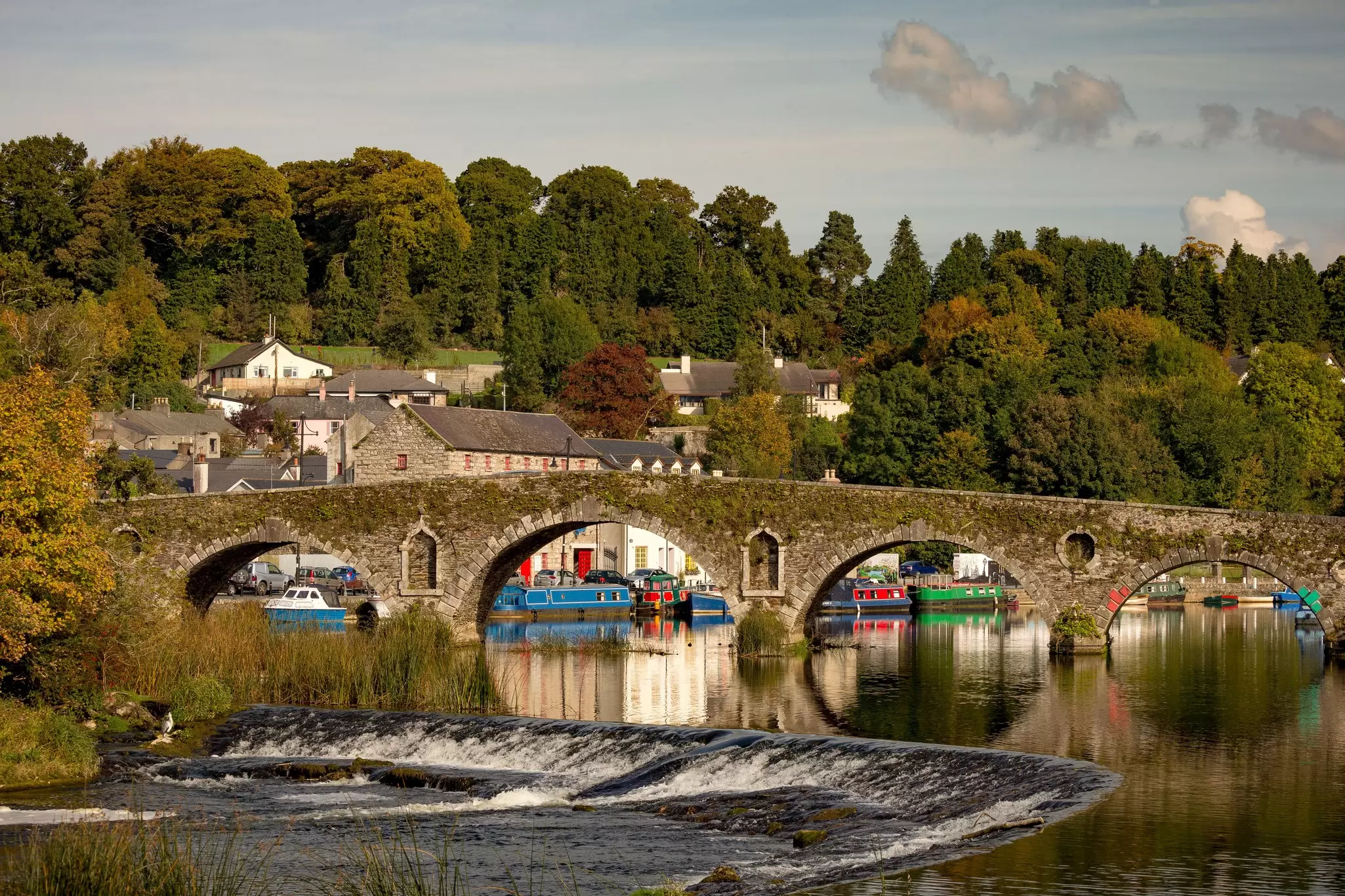 A riverside village of stone buildings with canal boats and a stone bridge 