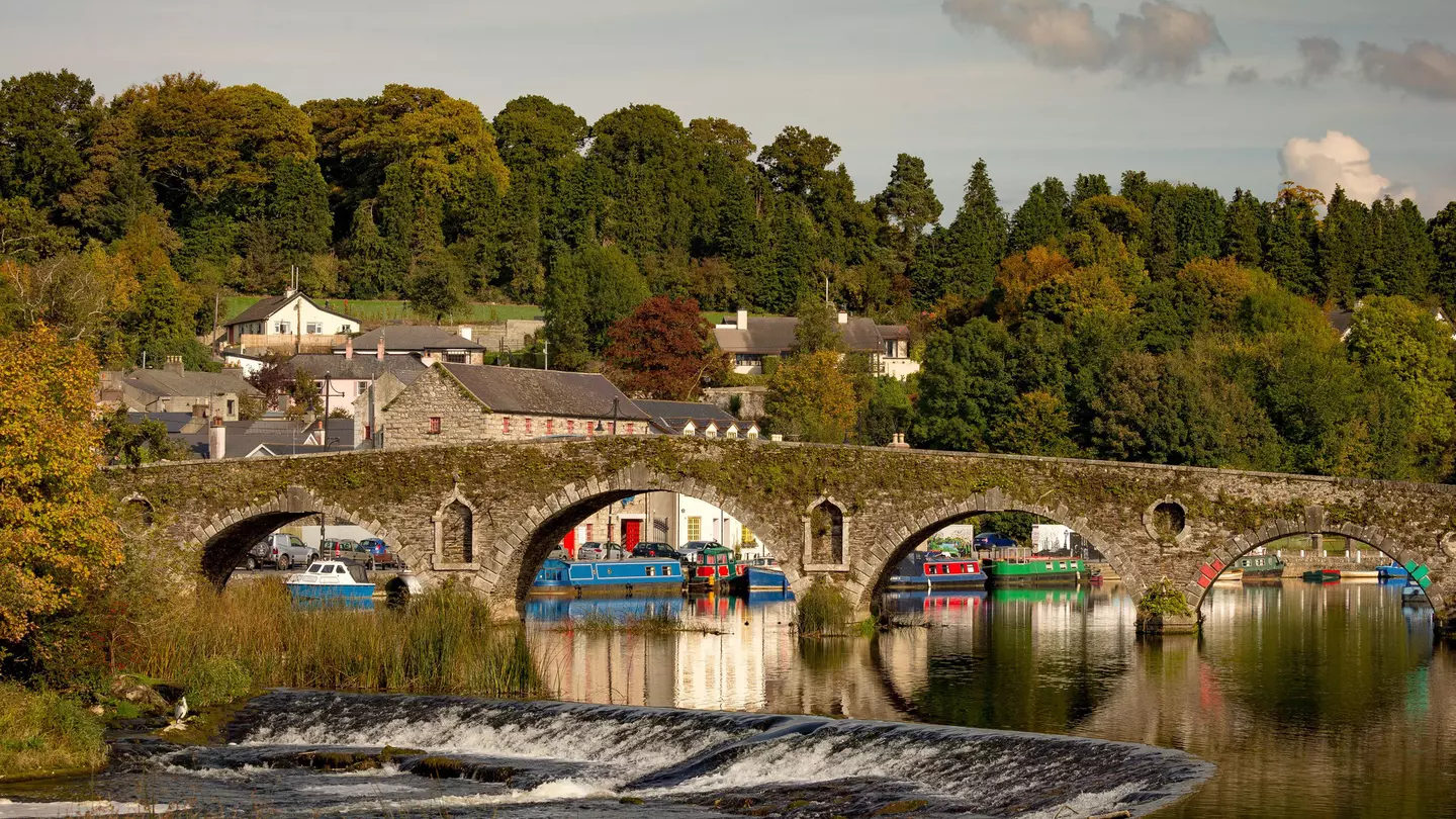 A riverside village of stone buildings with canal boats and a stone bridge 