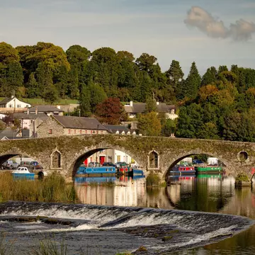 A riverside village of stone buildings with canal boats and a stone bridge 