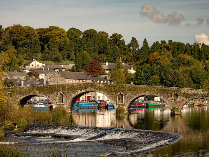 A riverside village of stone buildings with canal boats and a stone bridge 