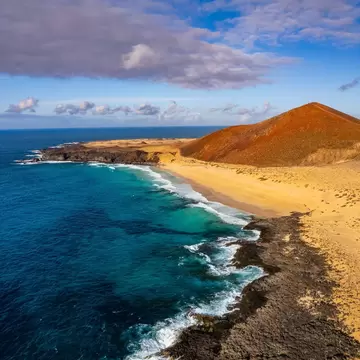 Turquoise surf meets a long golden beach below the red volcanic cone of Montaña Bermeja on La Graciosa