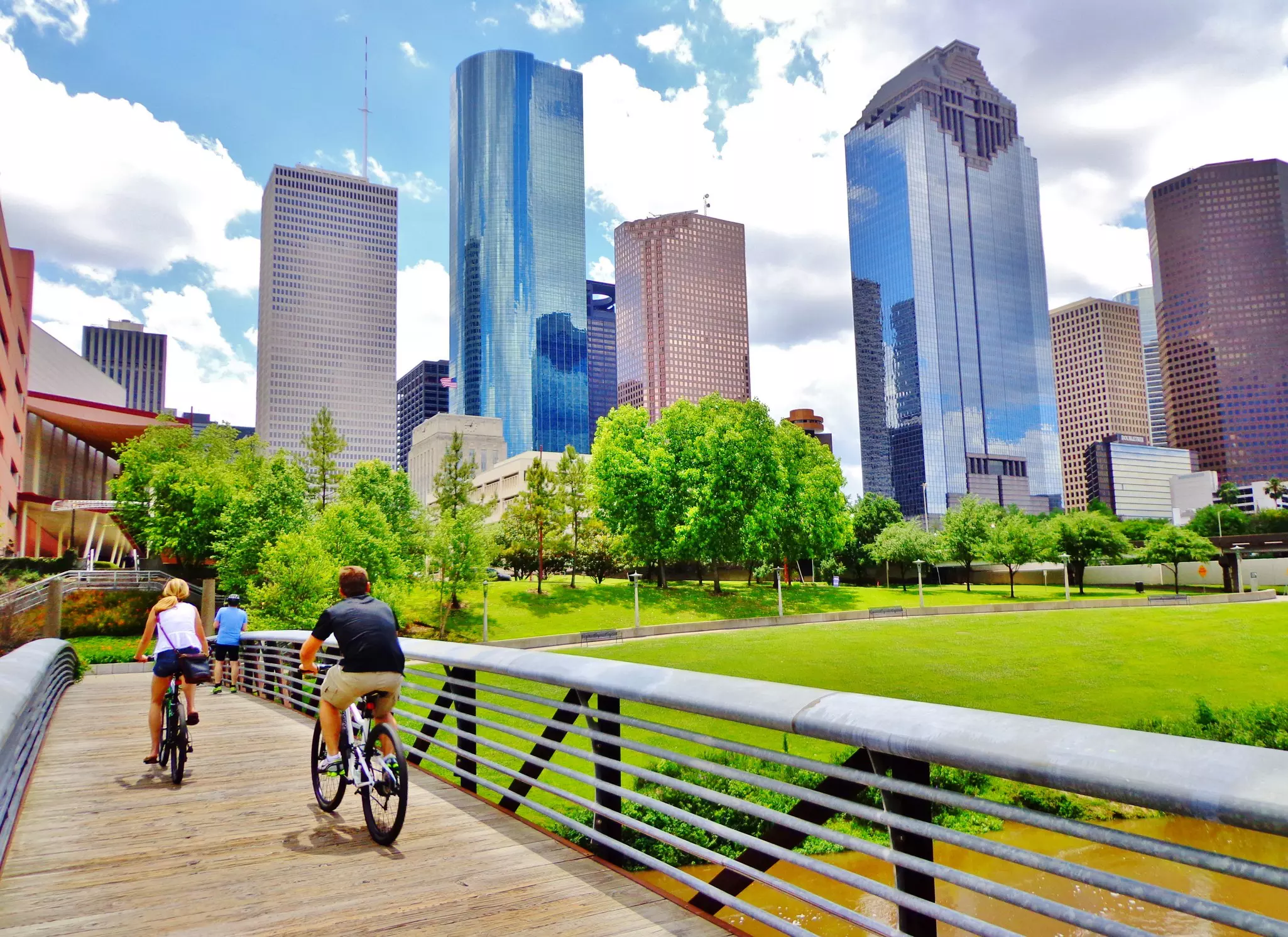Two cyclists ride bikes over a wooden bridge in parkland adjacent to high-rise city buildings.