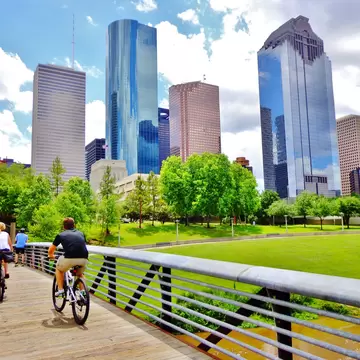 Two cyclists ride bikes over a wooden bridge in parkland adjacent to high-rise city buildings.