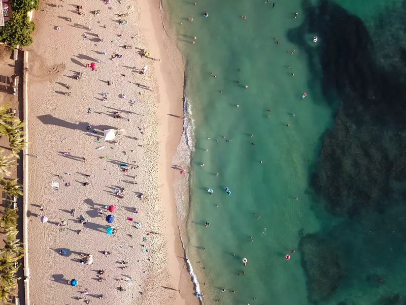 Overhead view of sandy beach with sunbathers and swimmers in the ocean to the right on a sunny day.