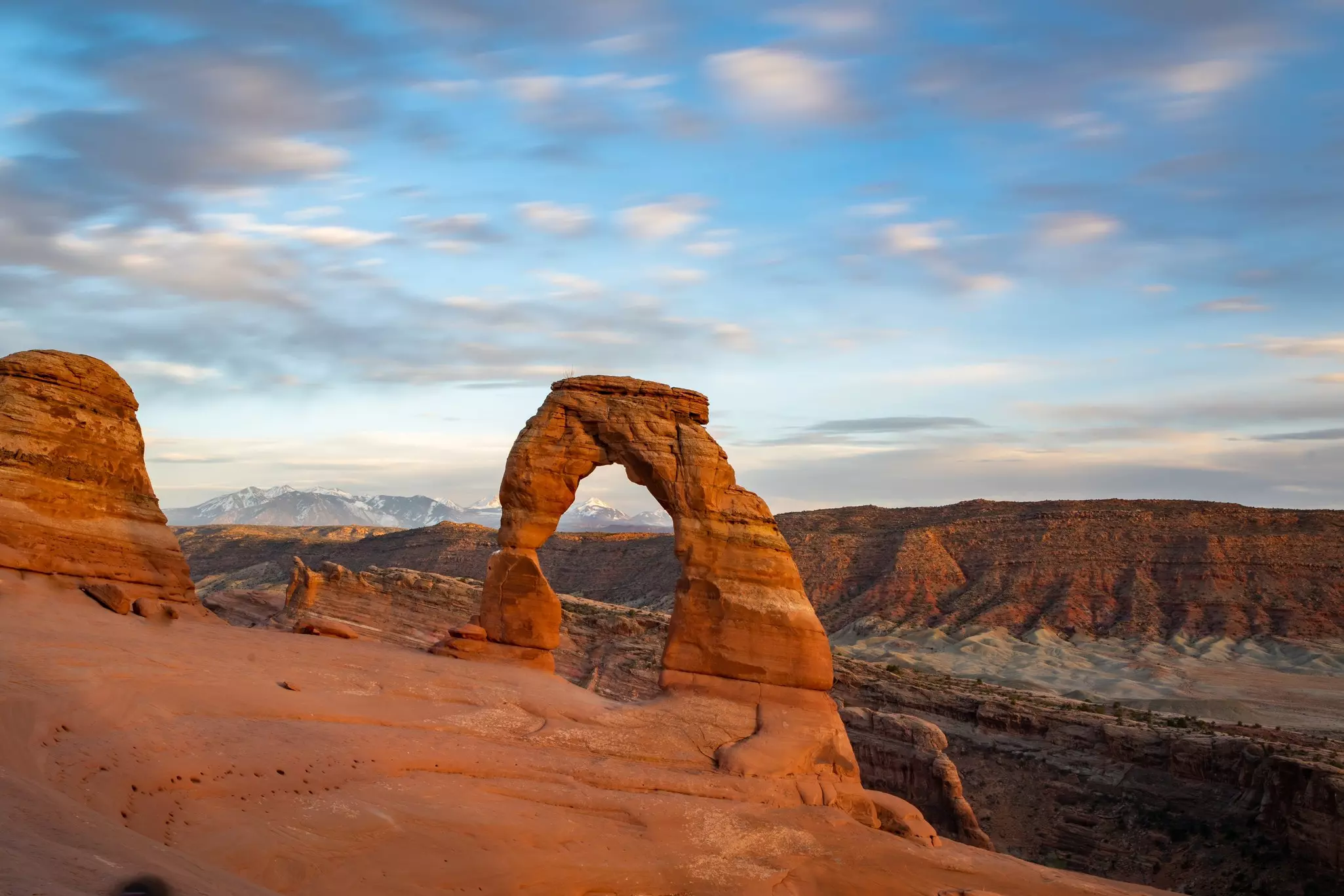 Wide shot of natural red rock arch with buttes in the distance and snow-covered peak in the far distance.