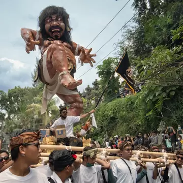 Men in white shirts and headcloths lifting a large painted demon statue on bamboo poles along a tree-lined street, Denpasar