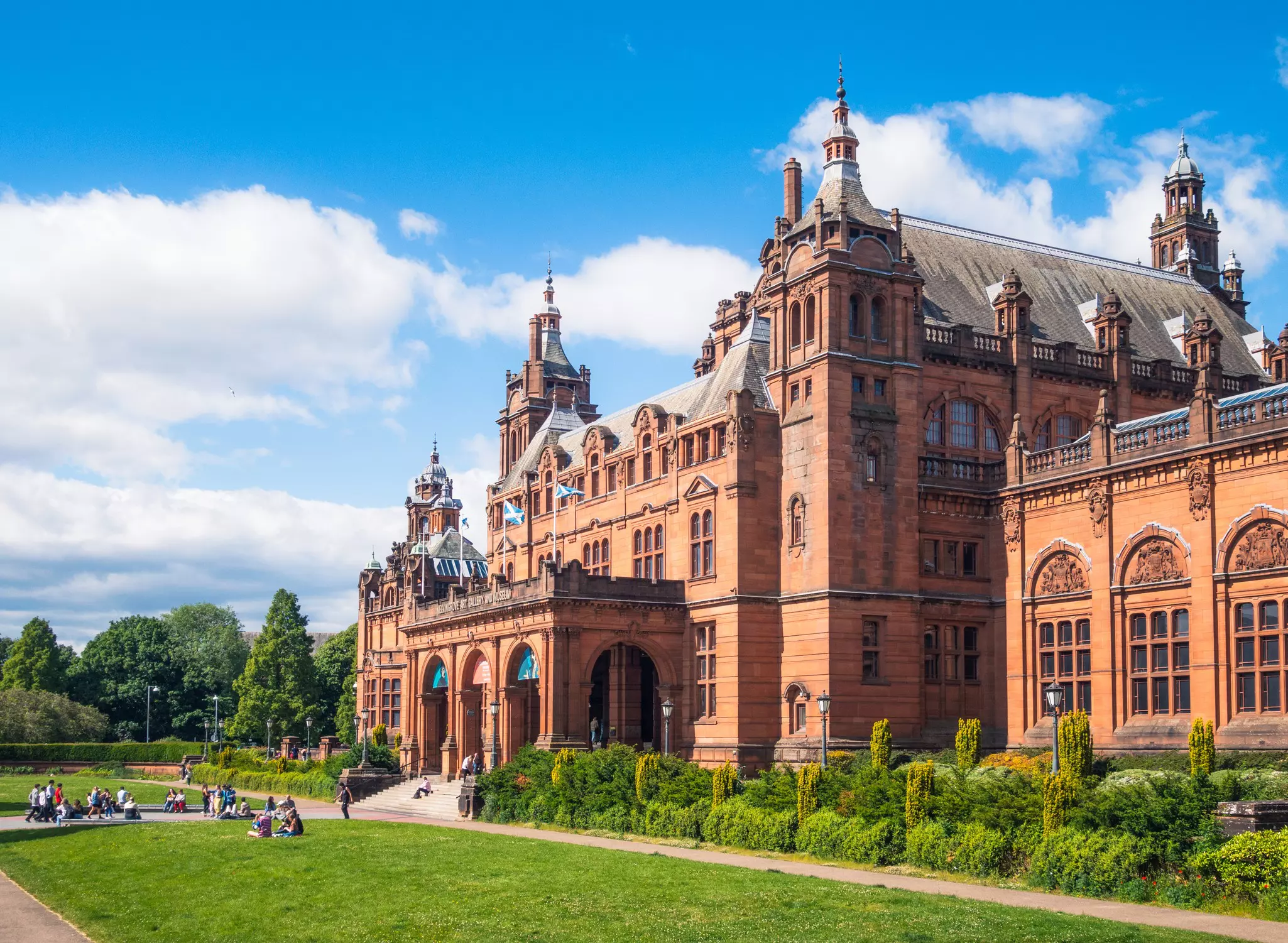 People are gathered on a green lawn and around the main entrance of a museum in Glasgow, Scotland, on a sunny day.