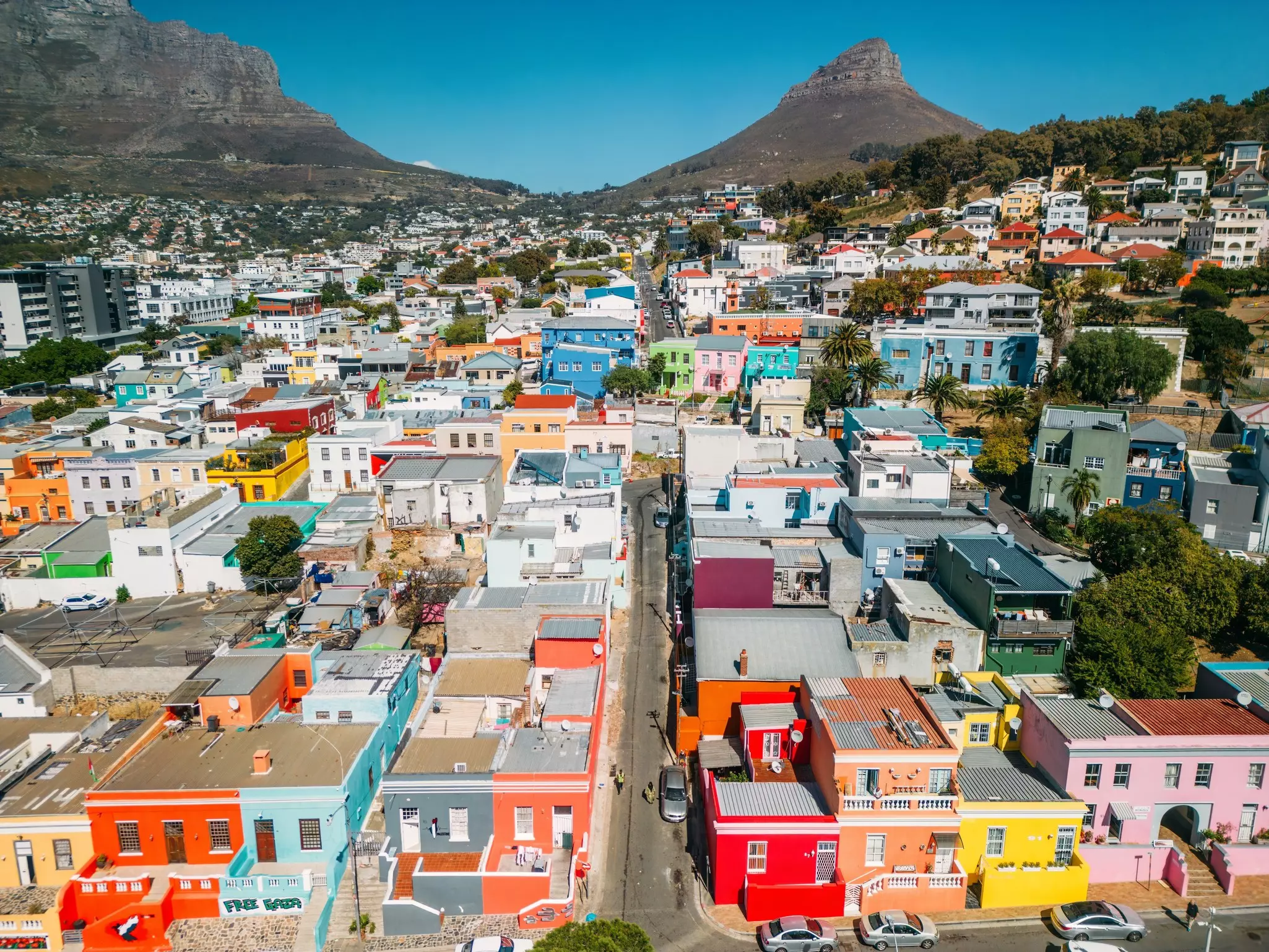 An aerial view of a city neighborhood with small, brightly colored buildings. A mountain with a rocky summit is visible in the distance.