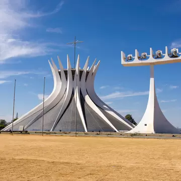 White curved concrete columns of Brasilia Cathedral beside a tall white bell tower, on dry grass under a blue sky.