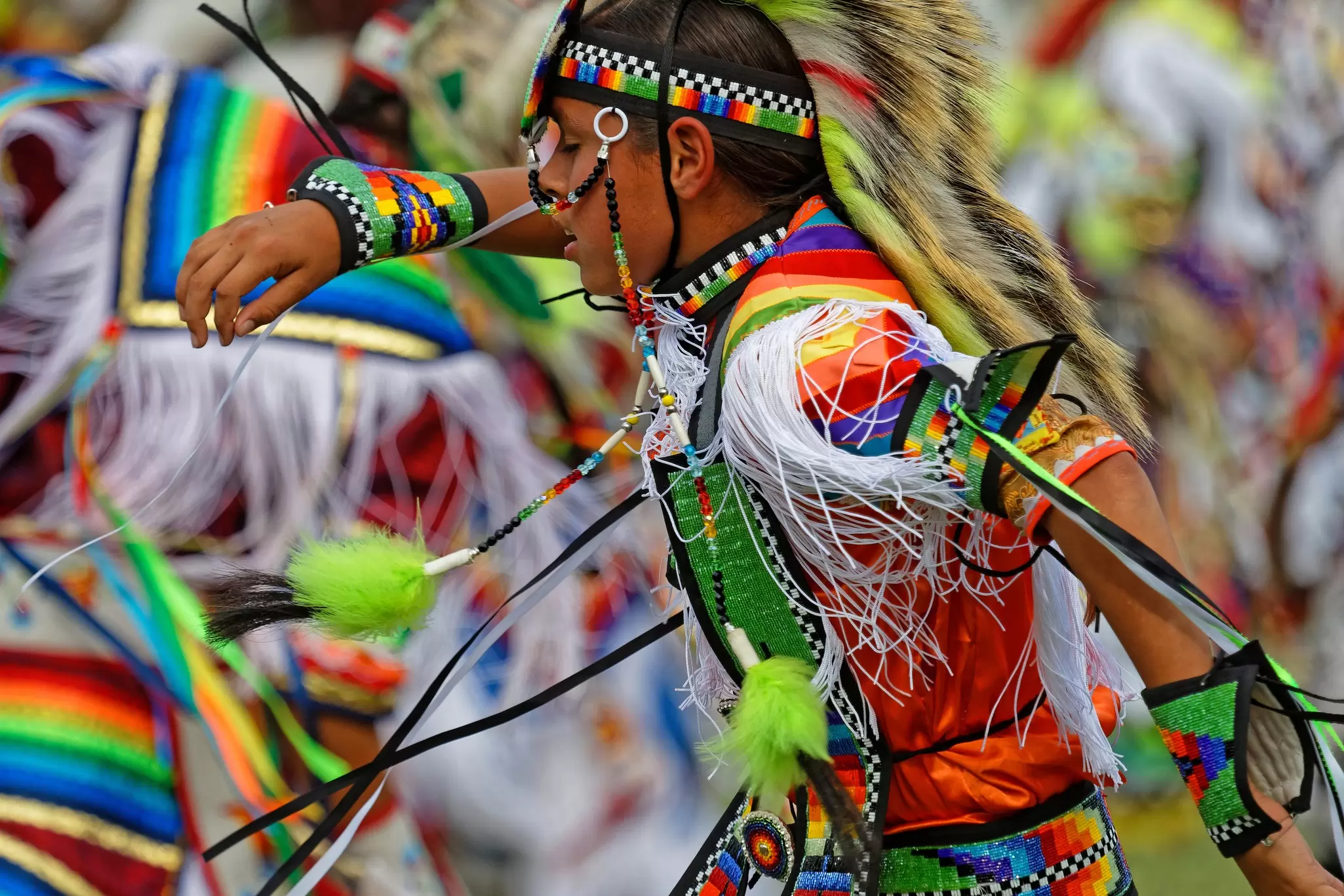 Young powwow dancer in vivid beaded rainbow regalia and a fur headdress performs in motion among other costumed dancers.