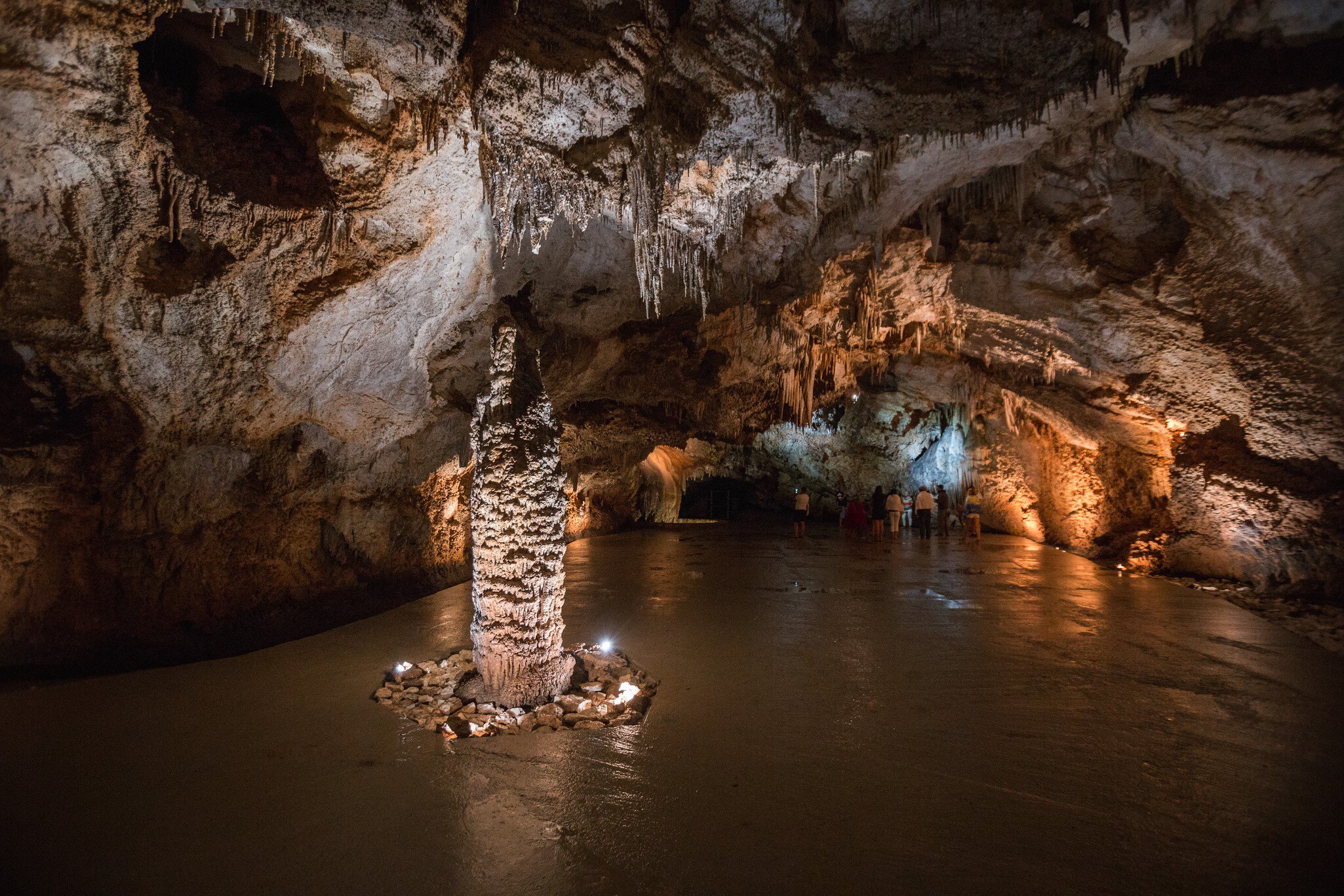 Tall knobbly stalagmite ringed by stones and small lights, with a group of visitors under a jagged rock ceiling beyond
