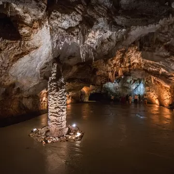 Tall knobbly stalagmite ringed by stones and small lights, with a group of visitors under a jagged rock ceiling beyond