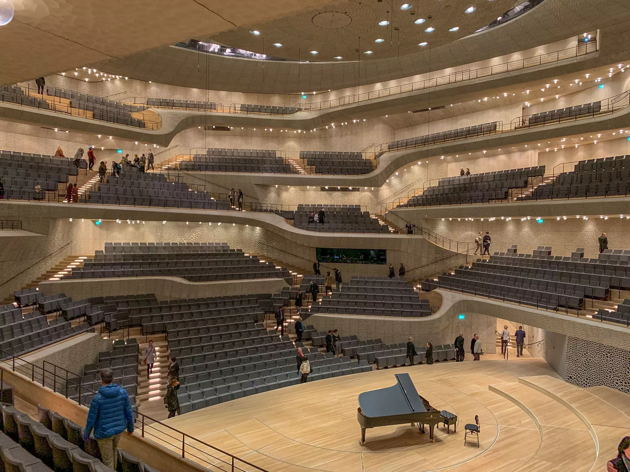 A concert grand piano is on the stage of an an ultra-modern concert hall with multiple balconies and tiers of seating.