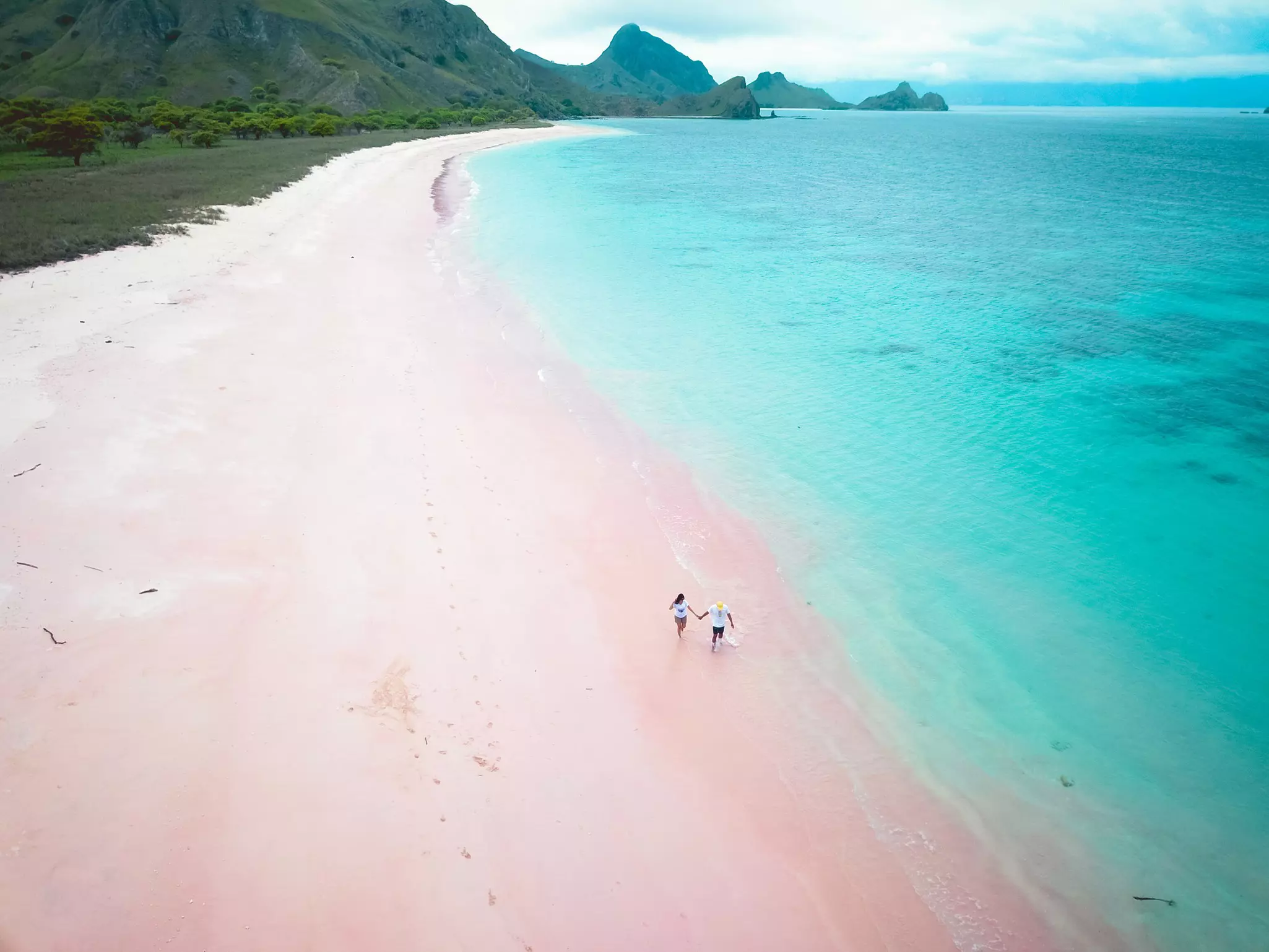 Aerial view of a couple walking and holding hands on a pink-sand beach with turquoise sea to the right and mountains in the distance.