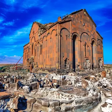 Ani Cathedral: roofless red and dark tufa church with tall blind arches, fallen masonry in front and hills under a blue sky