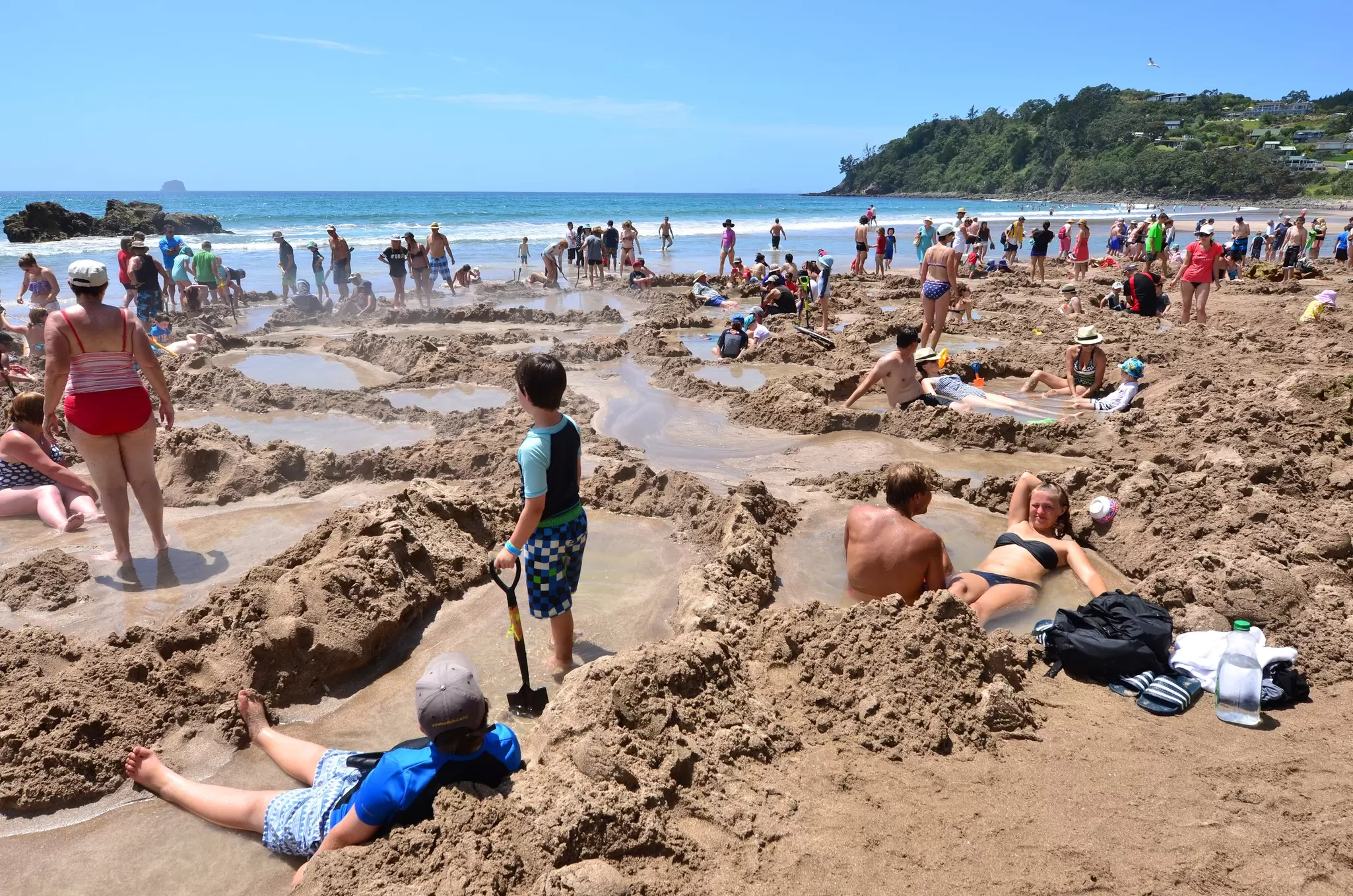 people soak in geothermal pools they've dug up on a beach in New Zealand on a sunny day