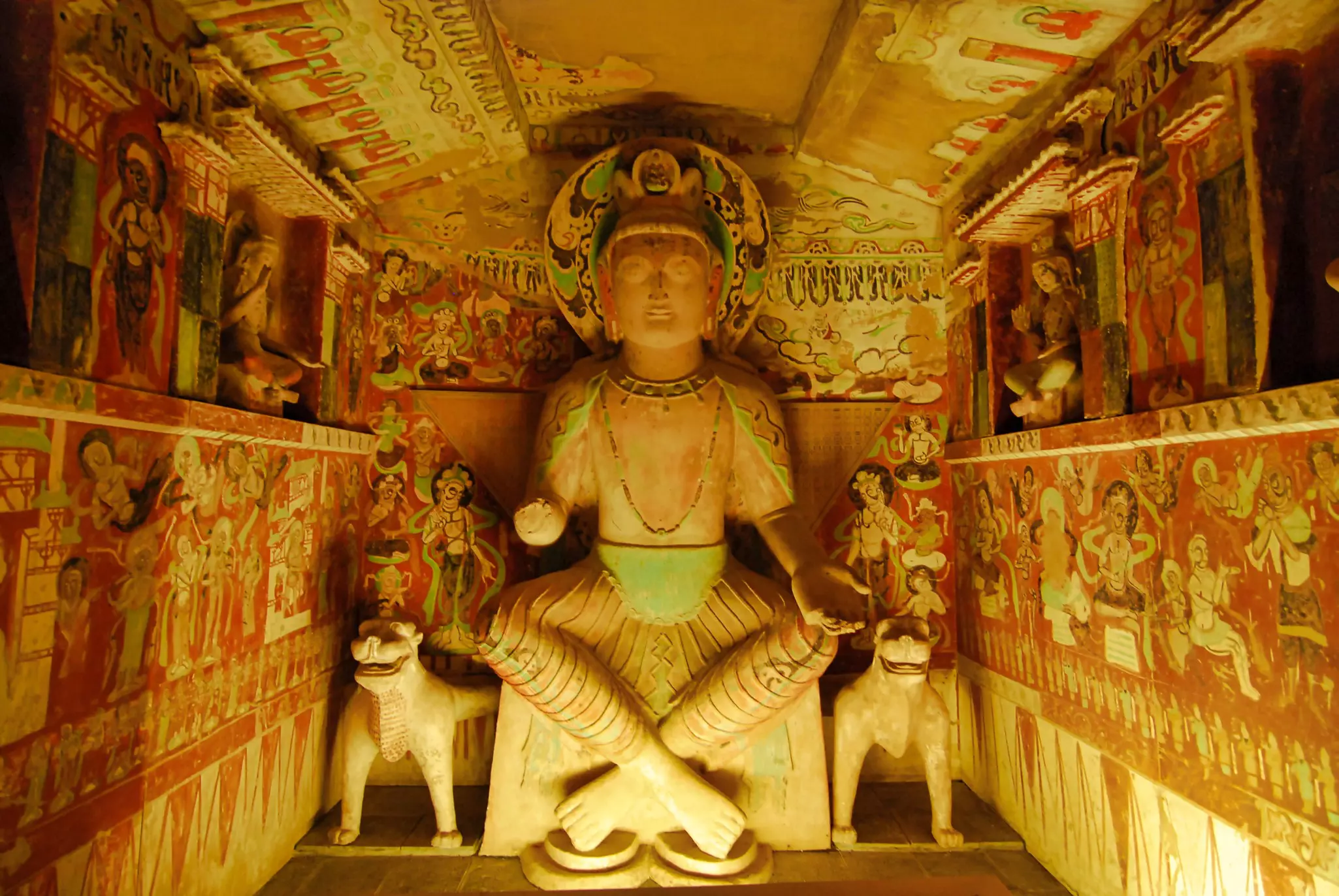 A statue of the Buddha is pictured in small cave with wall paintings depicting figures and other scenes.