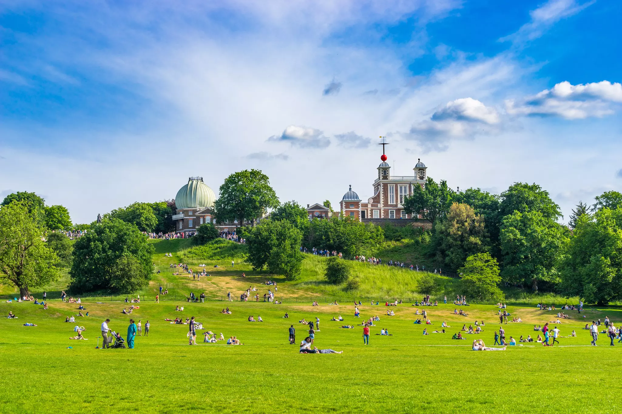Hilly park with people relaxing on wide grass lawn with trees, an observatory and a brick building in the distance on a sunny day.