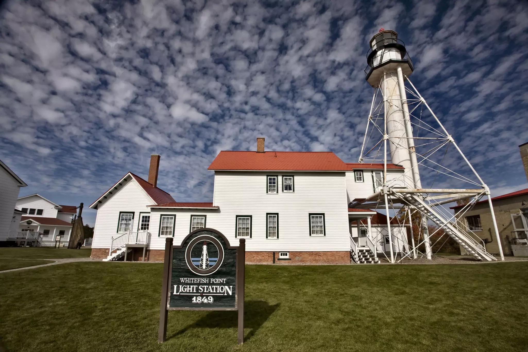 A lighthouse and red-roofed buildings. A sign reads Whitefish Point Light Station 1849.