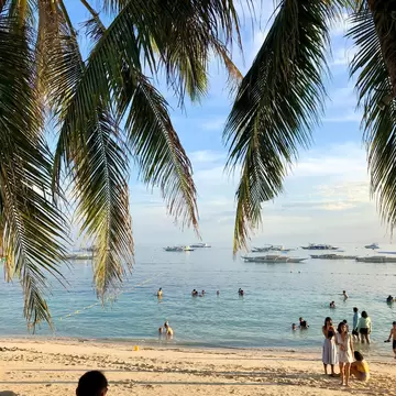Palm leaves over a sandy beach, people wading in calm turquoise water, outrigger boats offshore, Panglao, Philippines