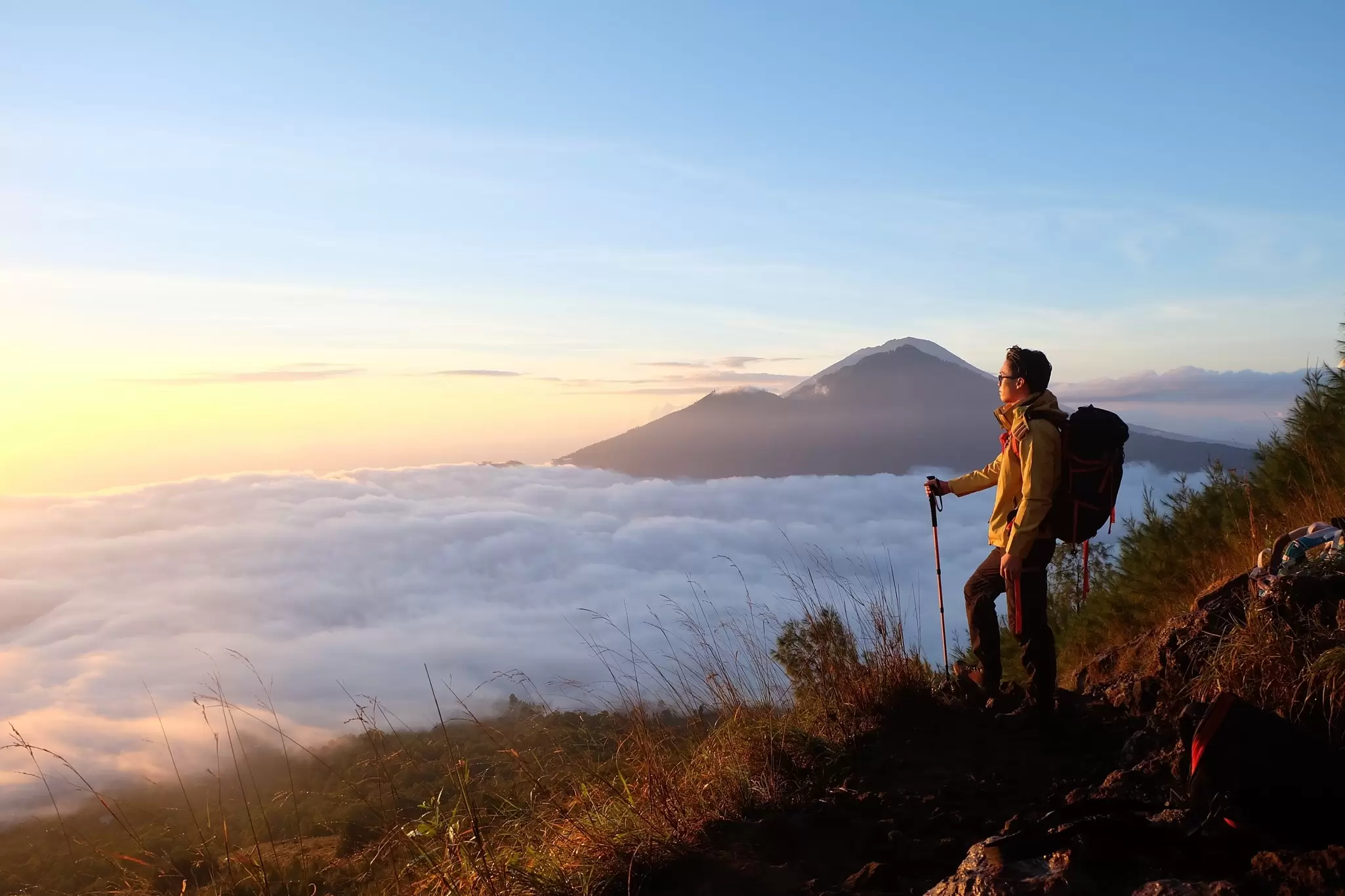 A person standing on a mountain peak with a backpack and hiking pole watching a sunrise