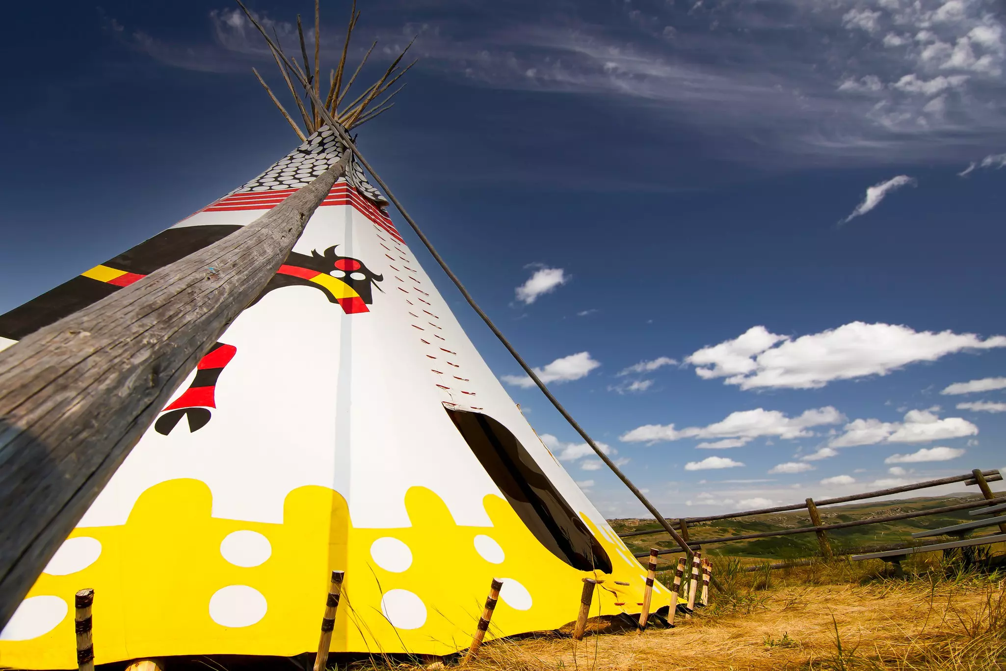 A colorful teepee, mostly white and yellow, set up in a field with blue sky and clouds in the distance.