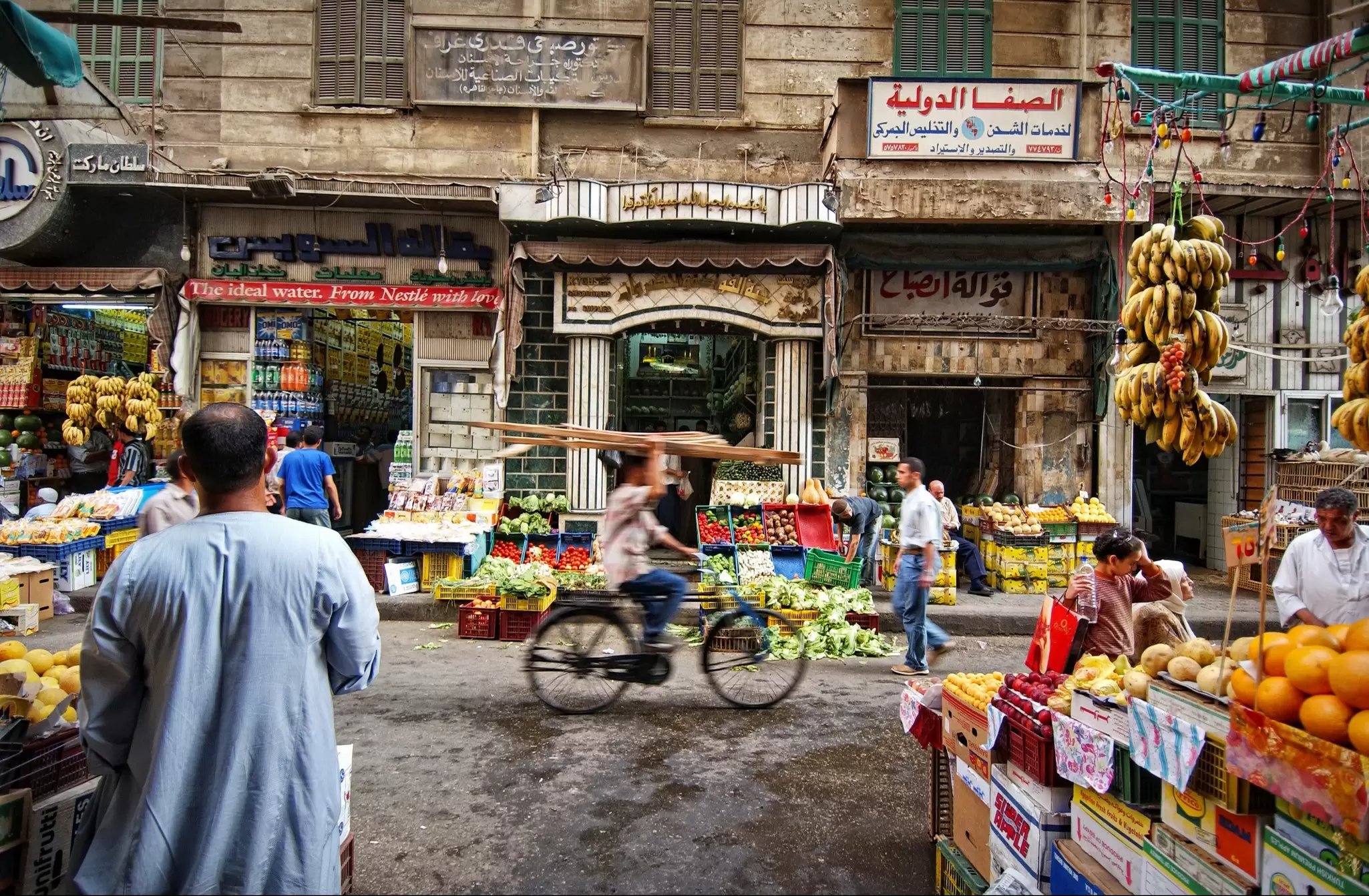 A man cycling down a narrow street with fruit and veg stalls on both sides.