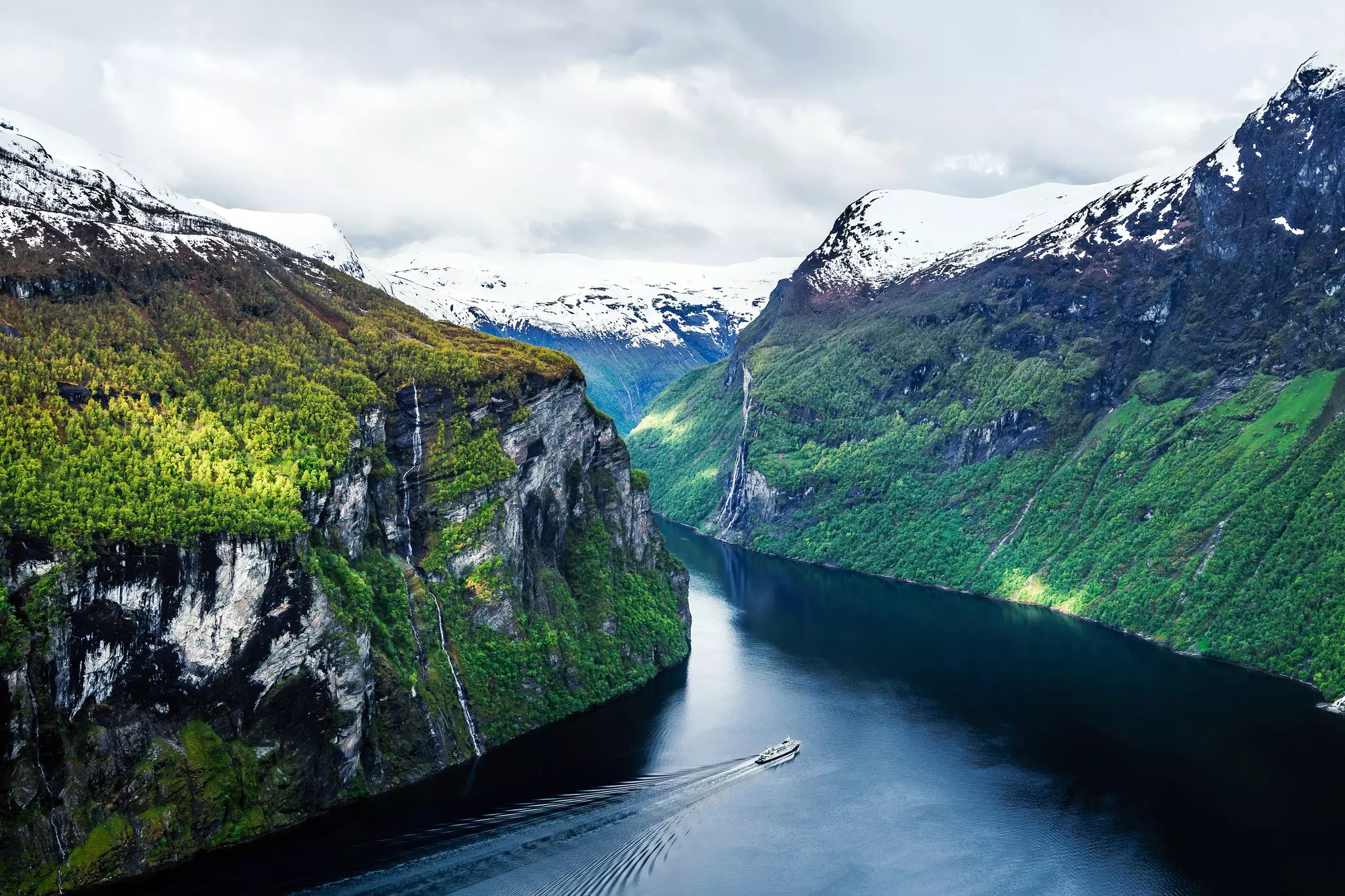 A wide view of a boat in a deep fjord. Lush cliffs plunge to the water, and snow-capped peaks are seen in the distance.