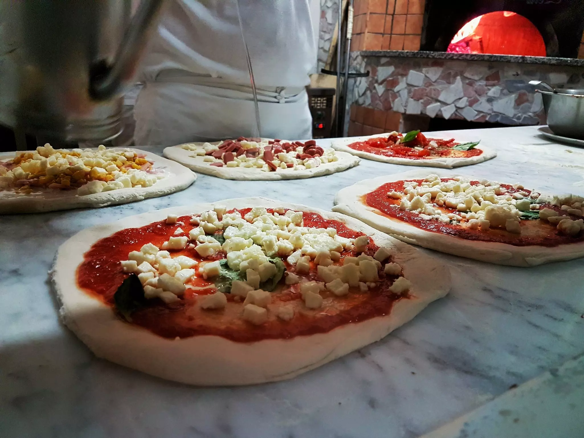 Five pizzas being prepared next to a wood-fired oven