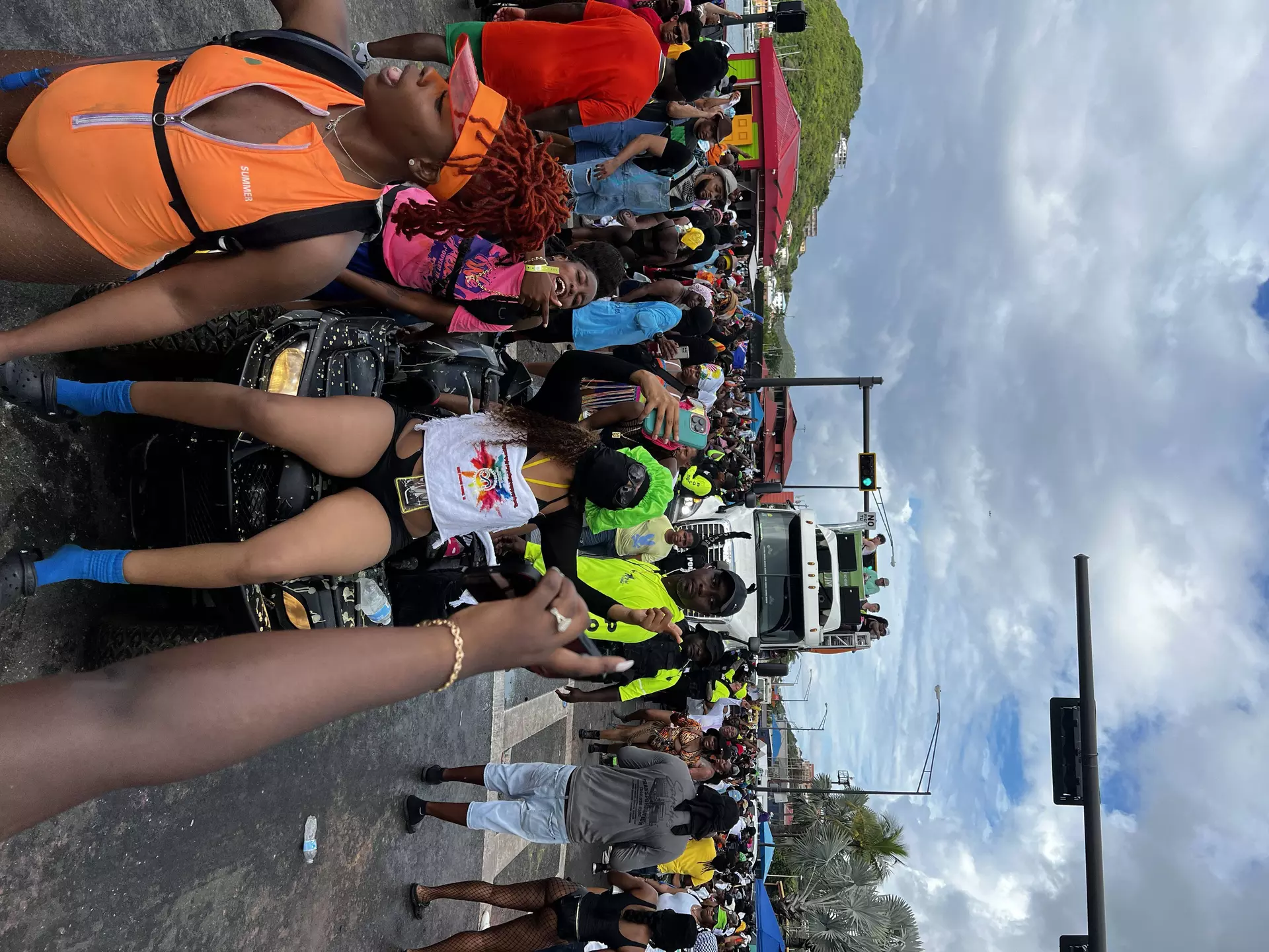 A woman poses on a small motorized cart while a large group of people walk pass during J'Ouvert in St Thomas. There is a large truck in the background.